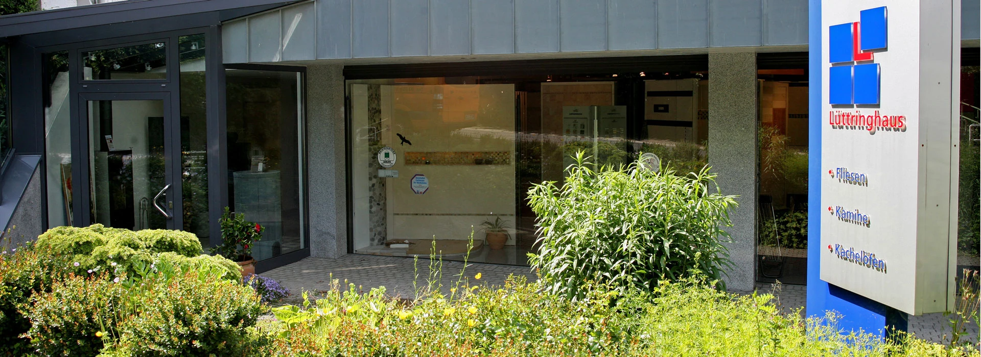 Eingang der Fliesenausstellung Lüttringhaus in der Hueckstraße in Lüdenscheid, davor Beete und ein Schild mit dem blau-roten Logo